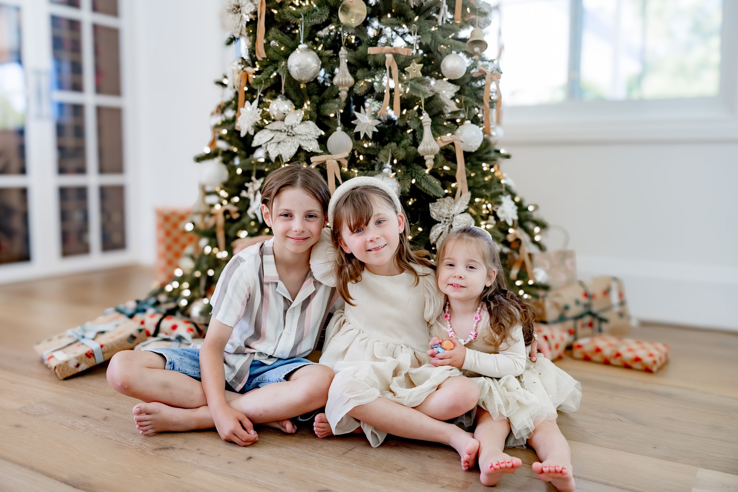 Children lying on wooden floor beside decorated Christmas tree in bright sunlit room.
