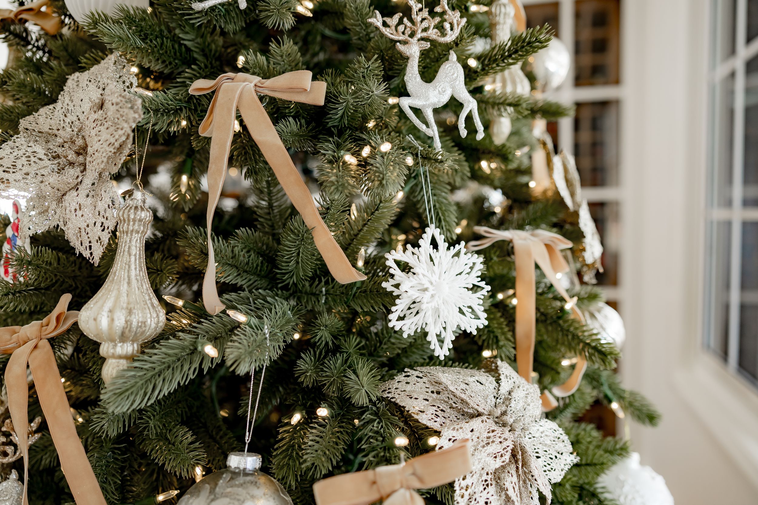 White snowflake ornaments and tan ribbons decorate a lush green Christmas tree next to a bright window.