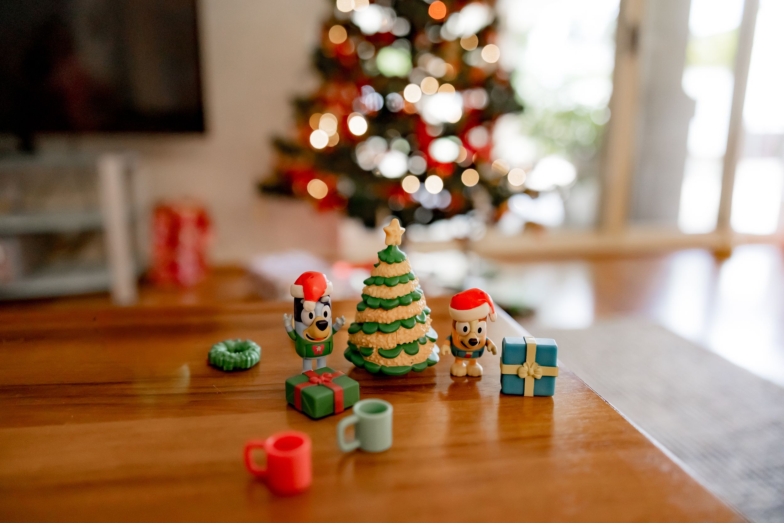 Small festive Christmas decorations and toys arranged on wooden table with decorated tree in background.