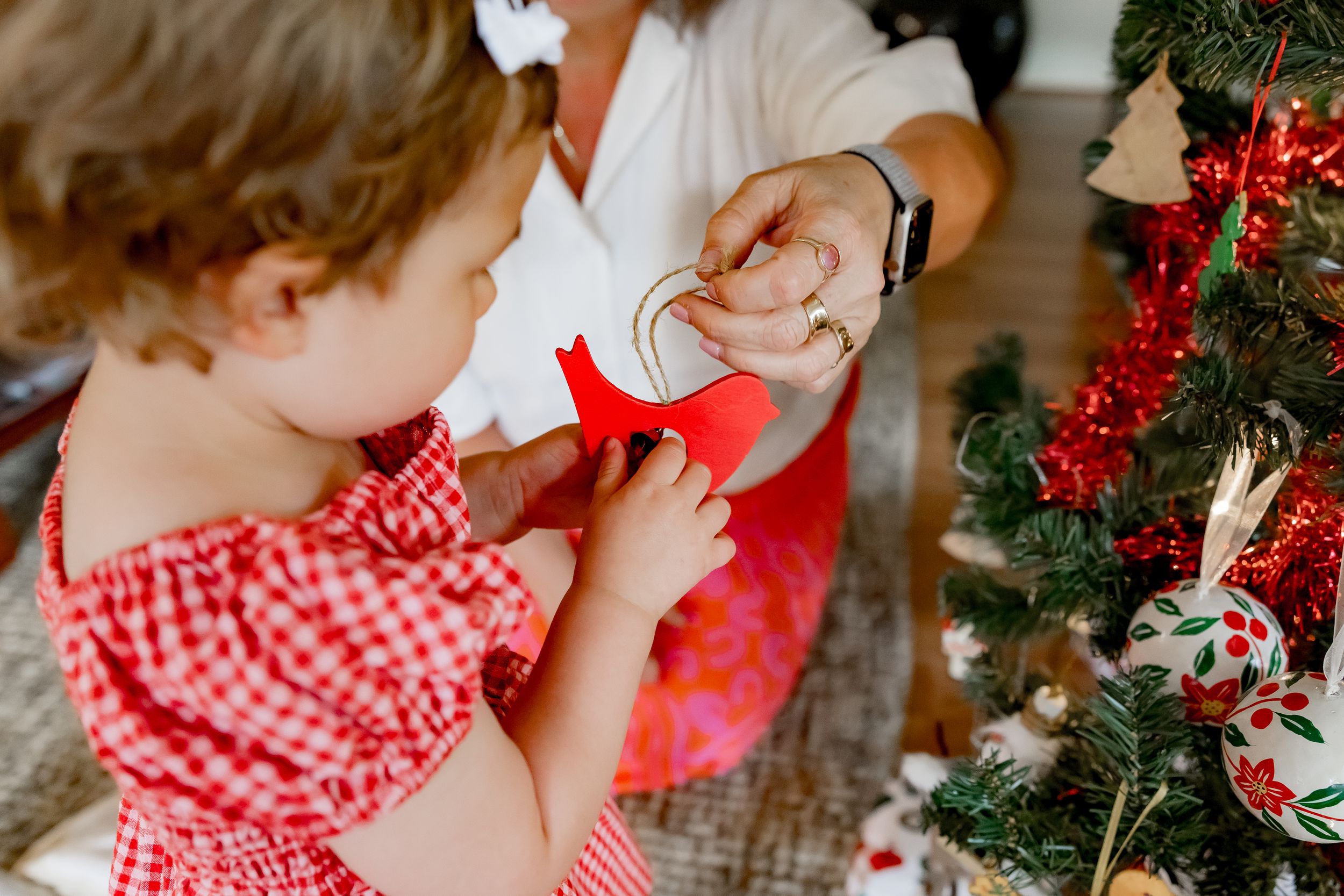 A child in a red gingham dress sits near a decorated Christmas tree with holiday ornaments and lights.