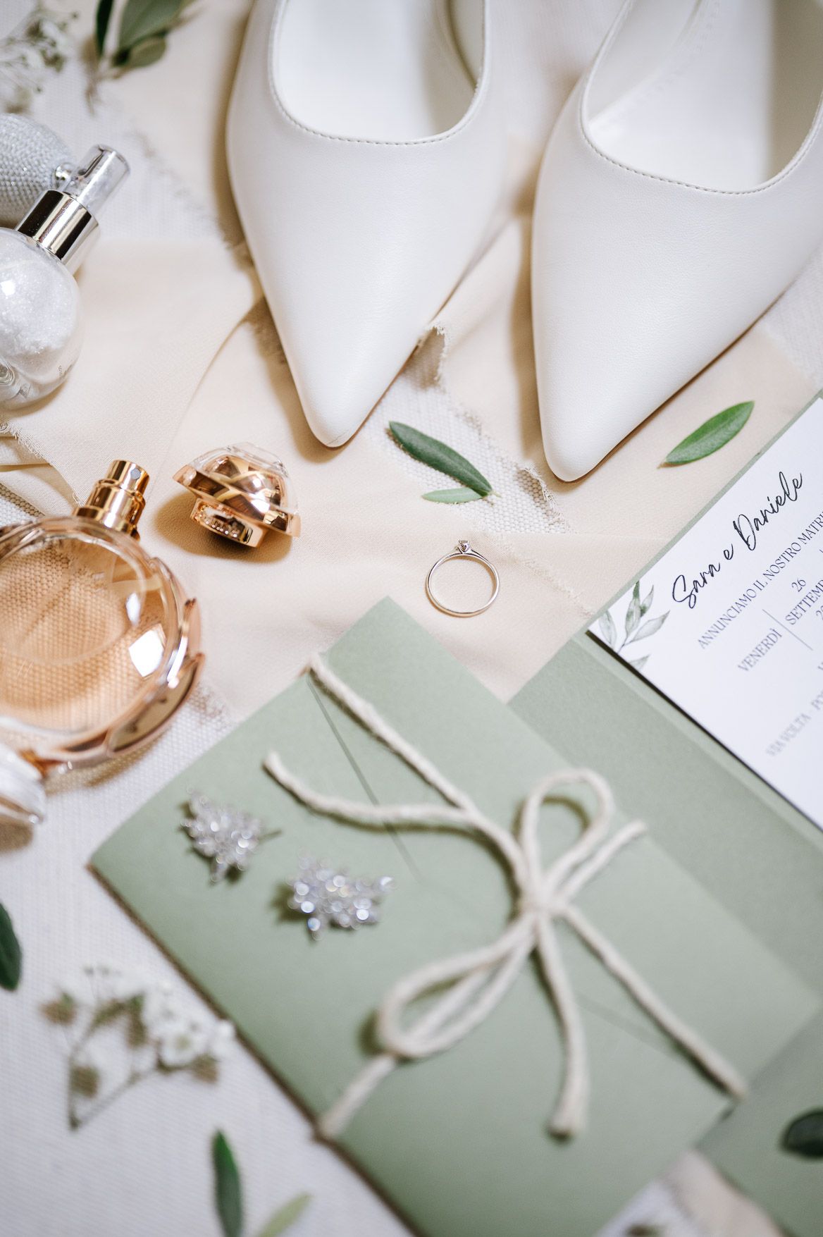 Elegant white bridal shoes and sage green wedding invitations with olive leaves and delicate ribbon details on cream background.