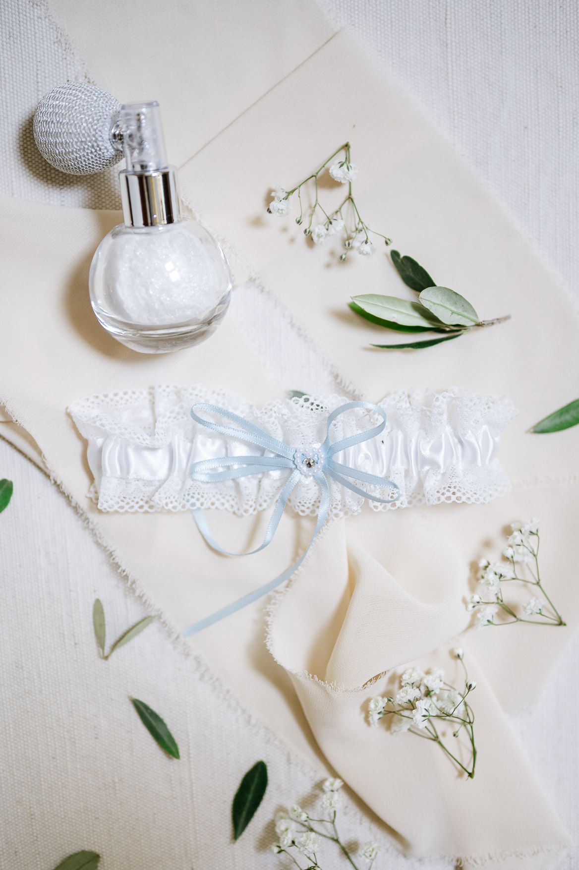 Elegant wedding accessories including white lace garter with blue bow, perfume bottle, and delicate baby's breath flowers.