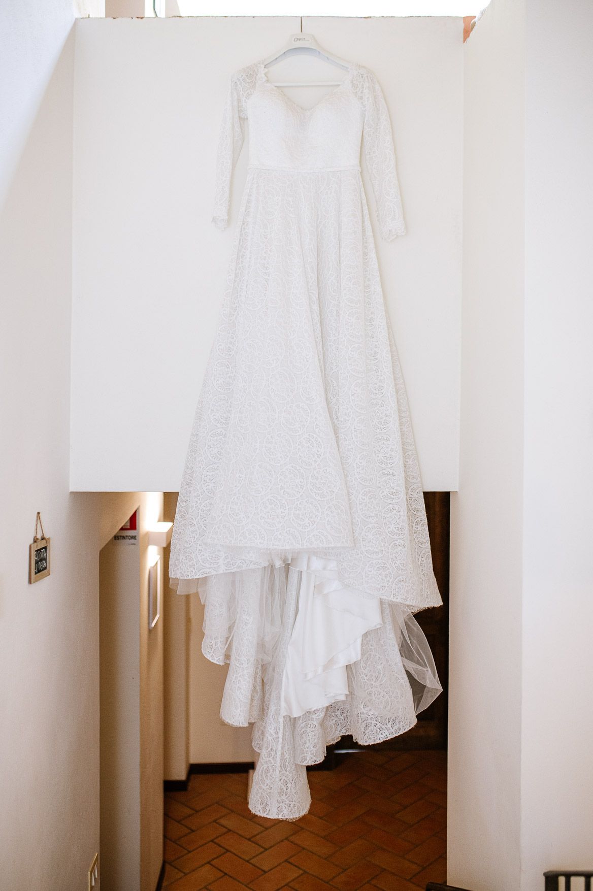 White vintage-style dress with lace details and flowing layers hangs elegantly against a light-colored wall.