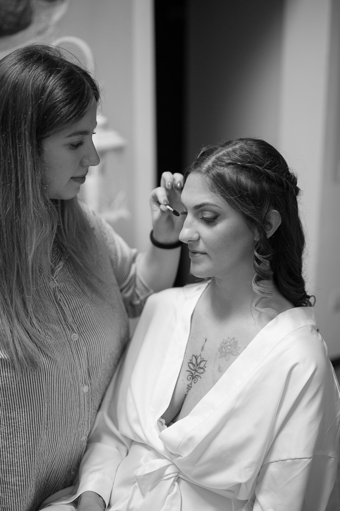 Black and white image showing intimate moments during bridal preparations with makeup application in a bright room.