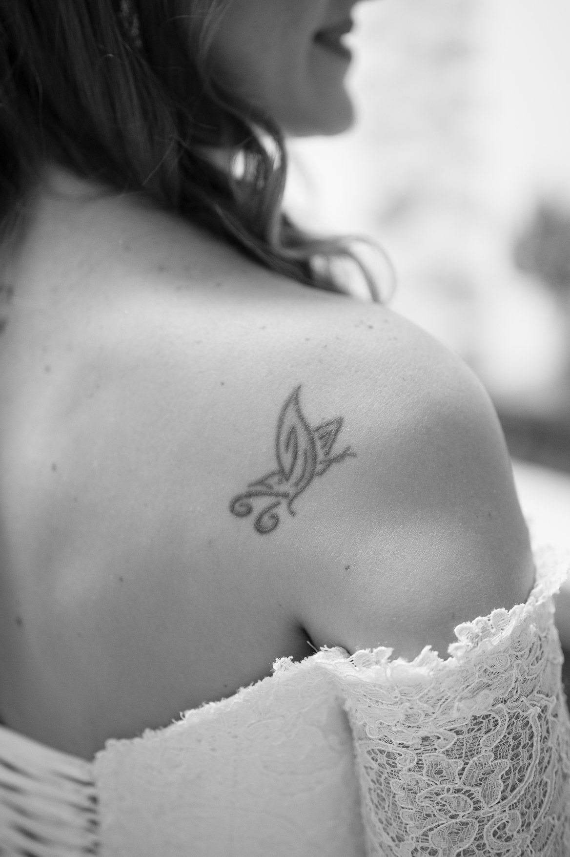 Black and white close-up of a delicate lace wedding dress detail against bare shoulders with a small butterfly tattoo.