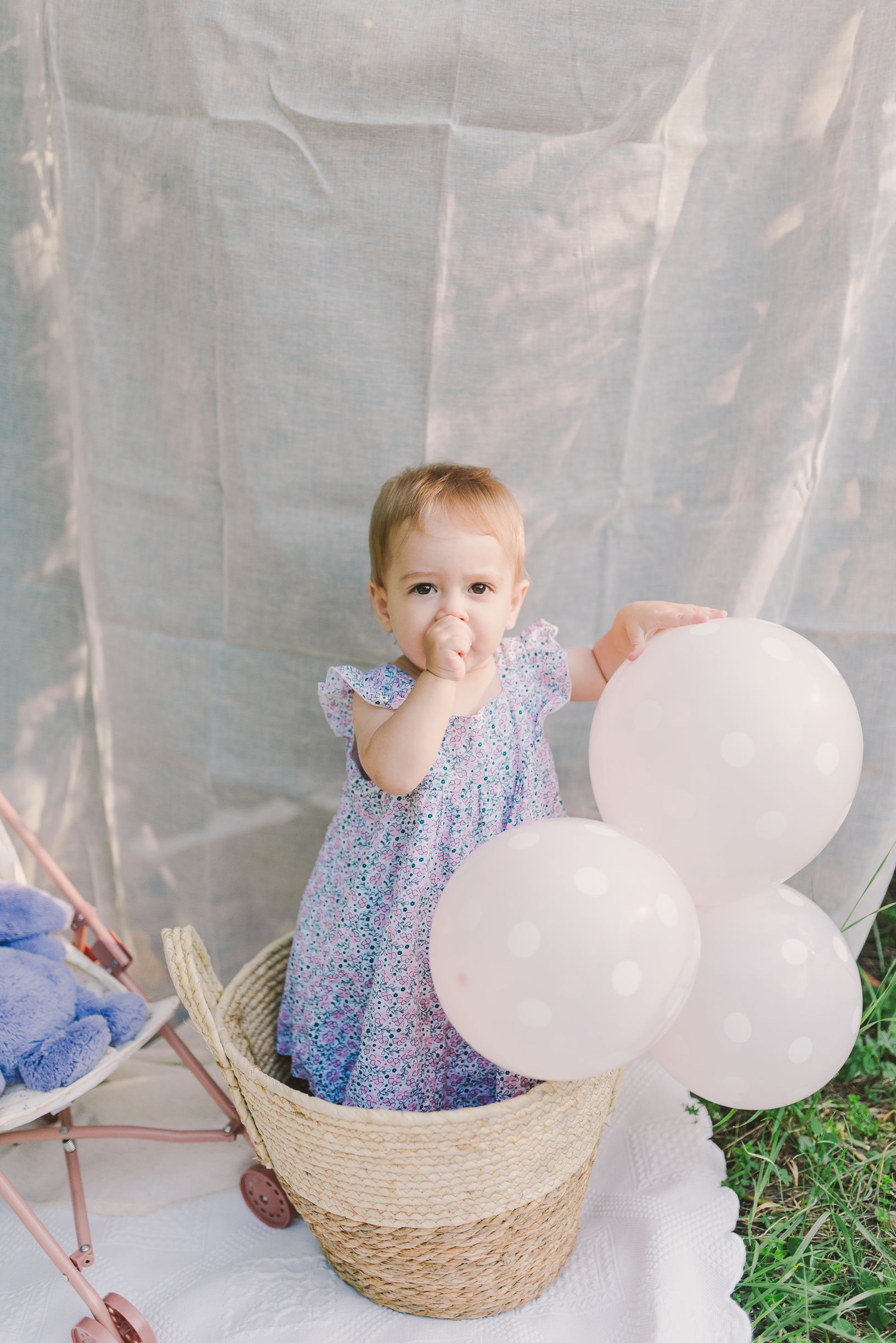 Child sitting in woven basket with white balloons against light backdrop.