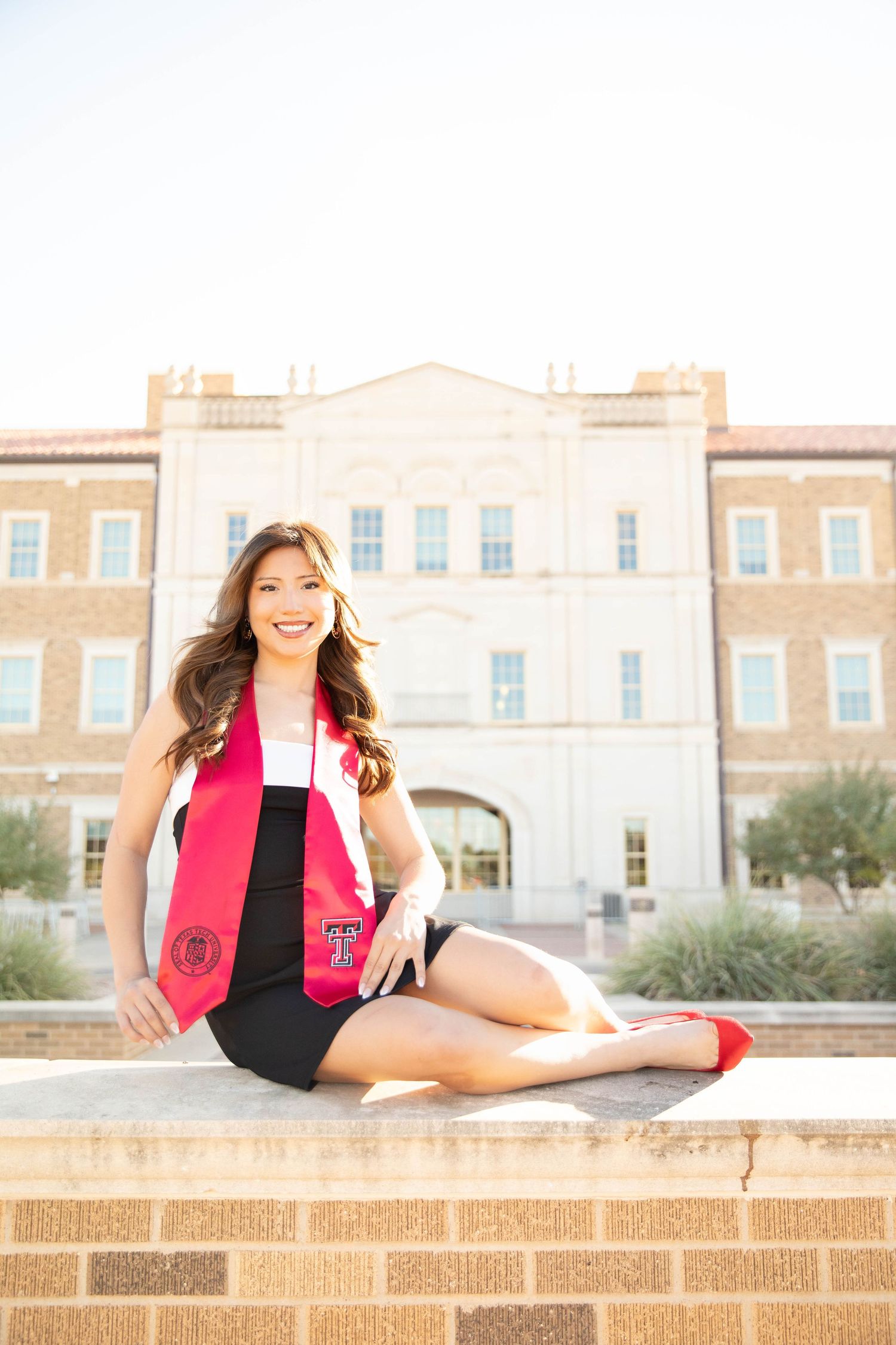 A person in a pink vest and black outfit poses on a brick ledge in front of a brick building.