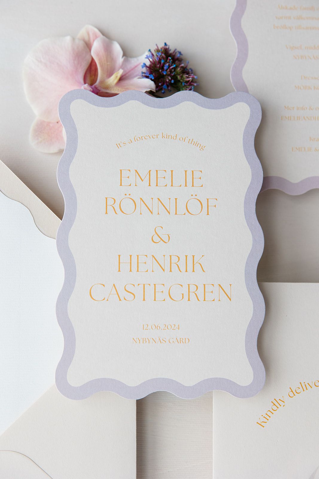 Elegant wedding invitation cards with lilac borders and gold text displayed on a light gray background.
