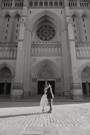 A romantic couple embraces in front of a grand Gothic cathedral with ornate stone architecture and rose window.