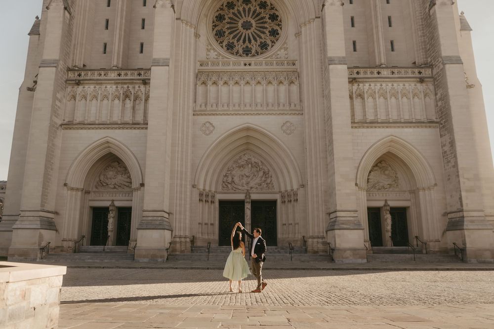 Two figures stand before the grand Gothic facade of a cathedral with ornate arched doorways and a rose window.