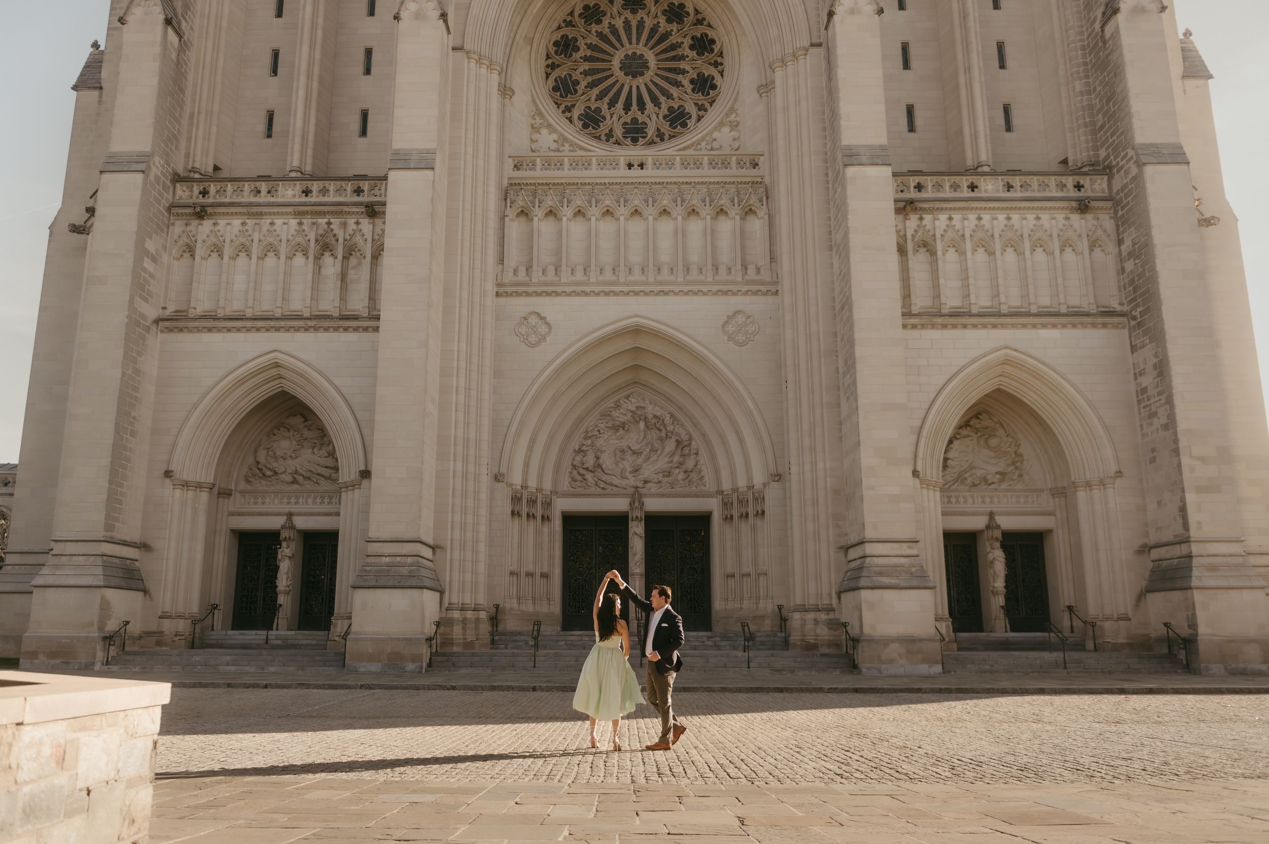 Two figures stand before the grand Gothic facade of a cathedral with ornate arched doorways and a rose window.