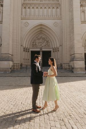 A couple in formal attire shares a romantic moment in front of a grand cathedral with ornate stone architecture.