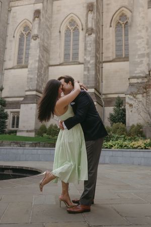 A romantic couple embraces in front of a gothic church with arched windows, the woman wearing a mint green dress.