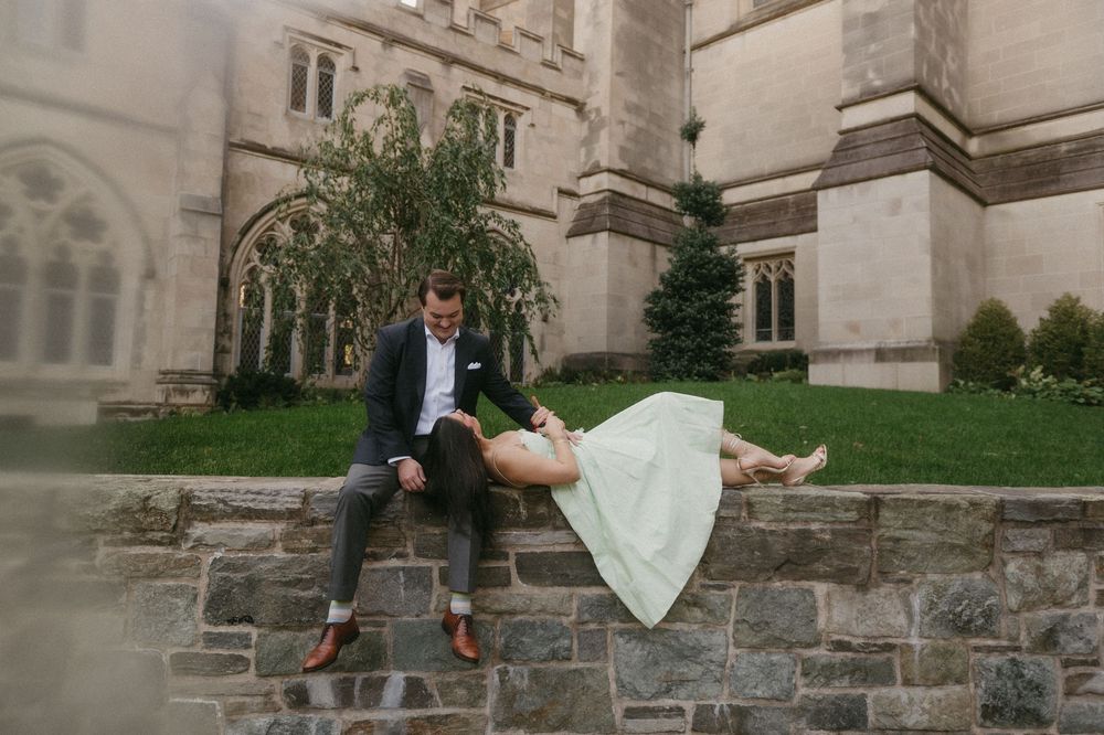 A romantic couple sits together on a stone wall outside a historic building on their wedding day.