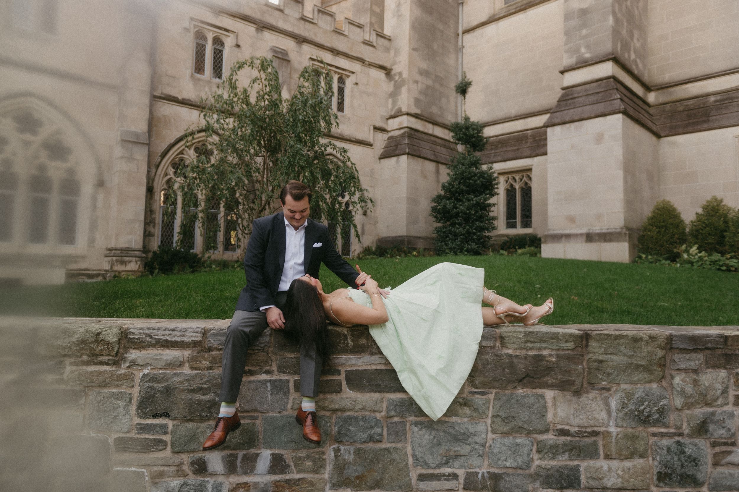 A romantic couple sits together on a stone wall outside a historic building on their wedding day.