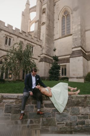 A couple in formal attire sits on a stone wall outside a gothic church in a playful levitation pose.