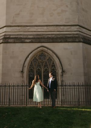 A couple stands outside a gothic church window with ornate stonework and iron fencing on a sunny day.