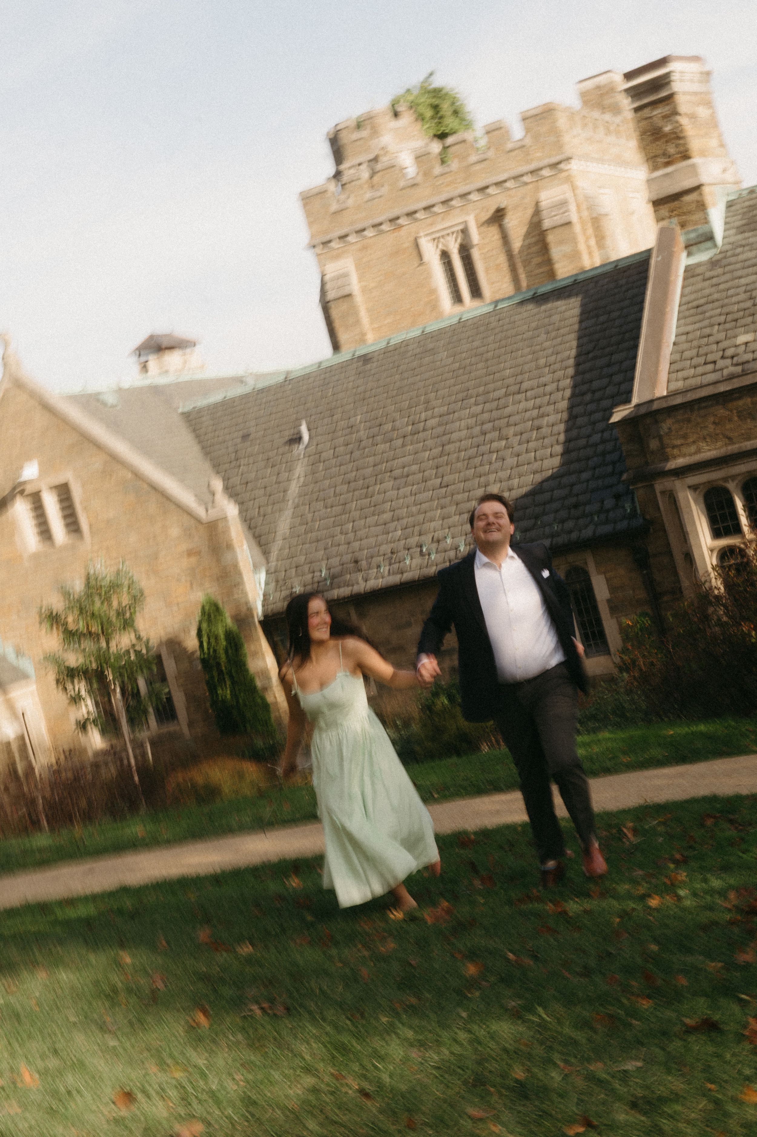 A wedding couple playfully runs and dances on the lawn in front of a stone castle-like building at sunset.