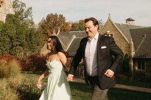 A couple in wedding attire holds hands while walking through a rustic outdoor venue with buildings in the background.