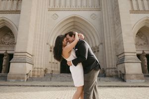 A romantic couple embraces and dips in front of an ornate stone cathedral with arched architectural details.