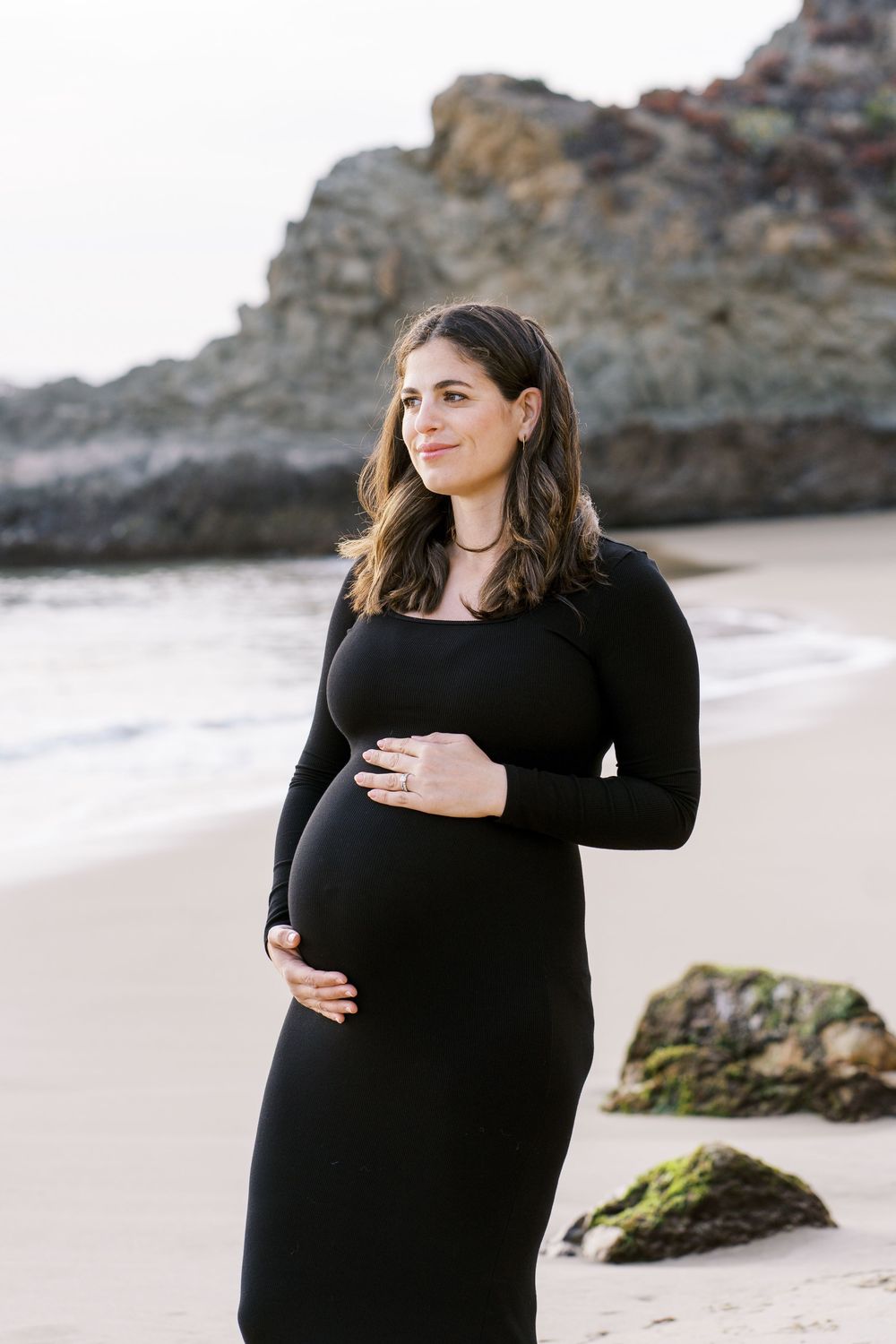A pregnant woman in a black dress stands on a beach near rocky cliffs during a maternity photoshoot.
