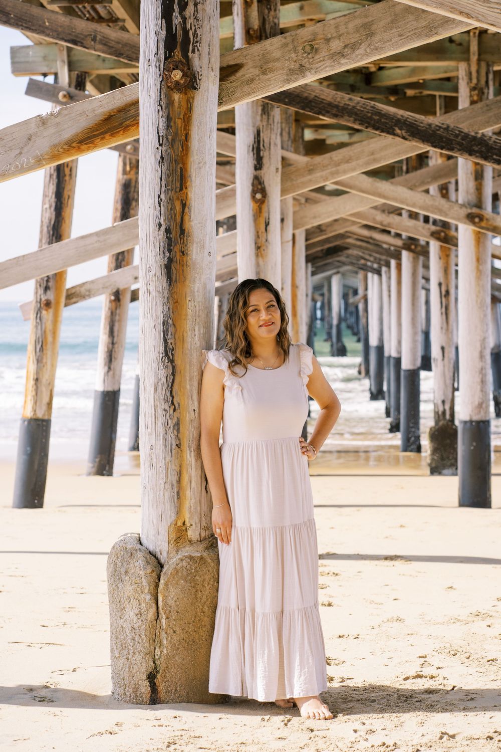 Woman in white summer dress poses under wooden pier at beach during sunny day.