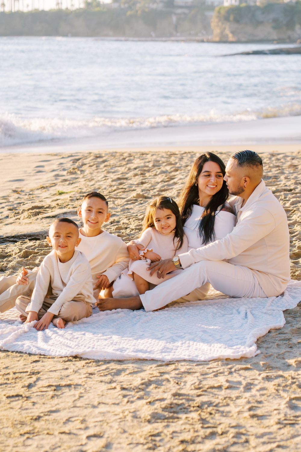 Family wearing white outfits relaxing together on a blanket at the beach during sunset.