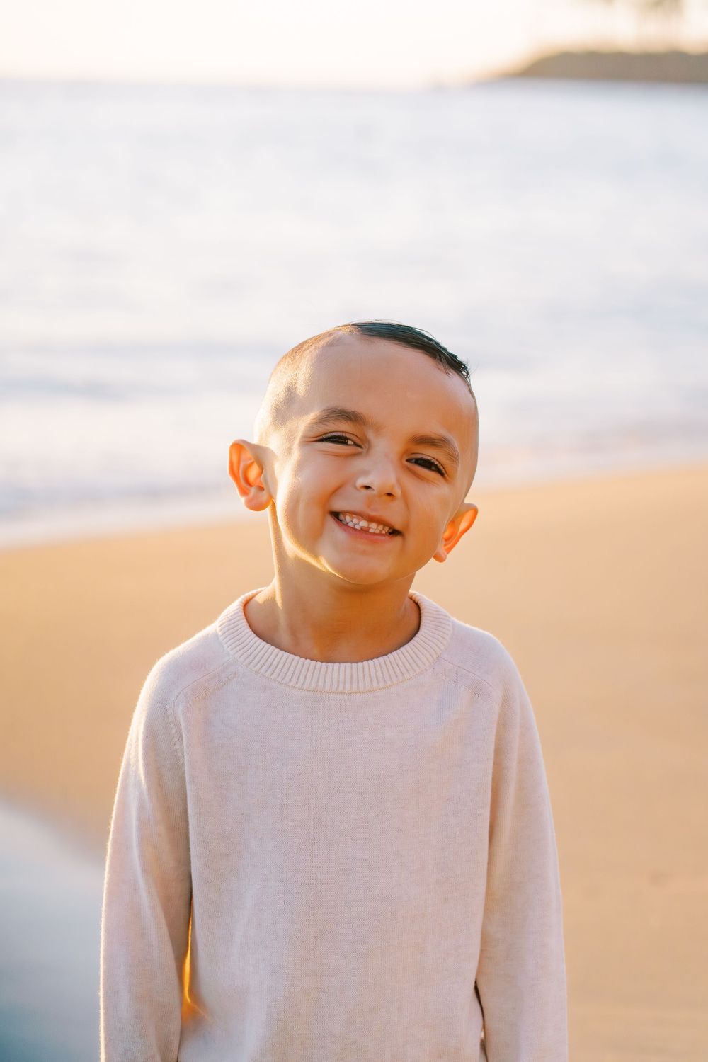 A young person in a white sweater smiles warmly during a golden hour photoshoot at the beach.
