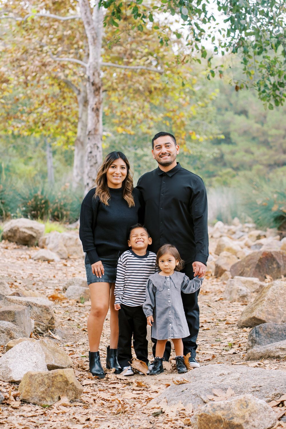 A family stands together on a rocky autumn trail surrounded by fall foliage and trees.