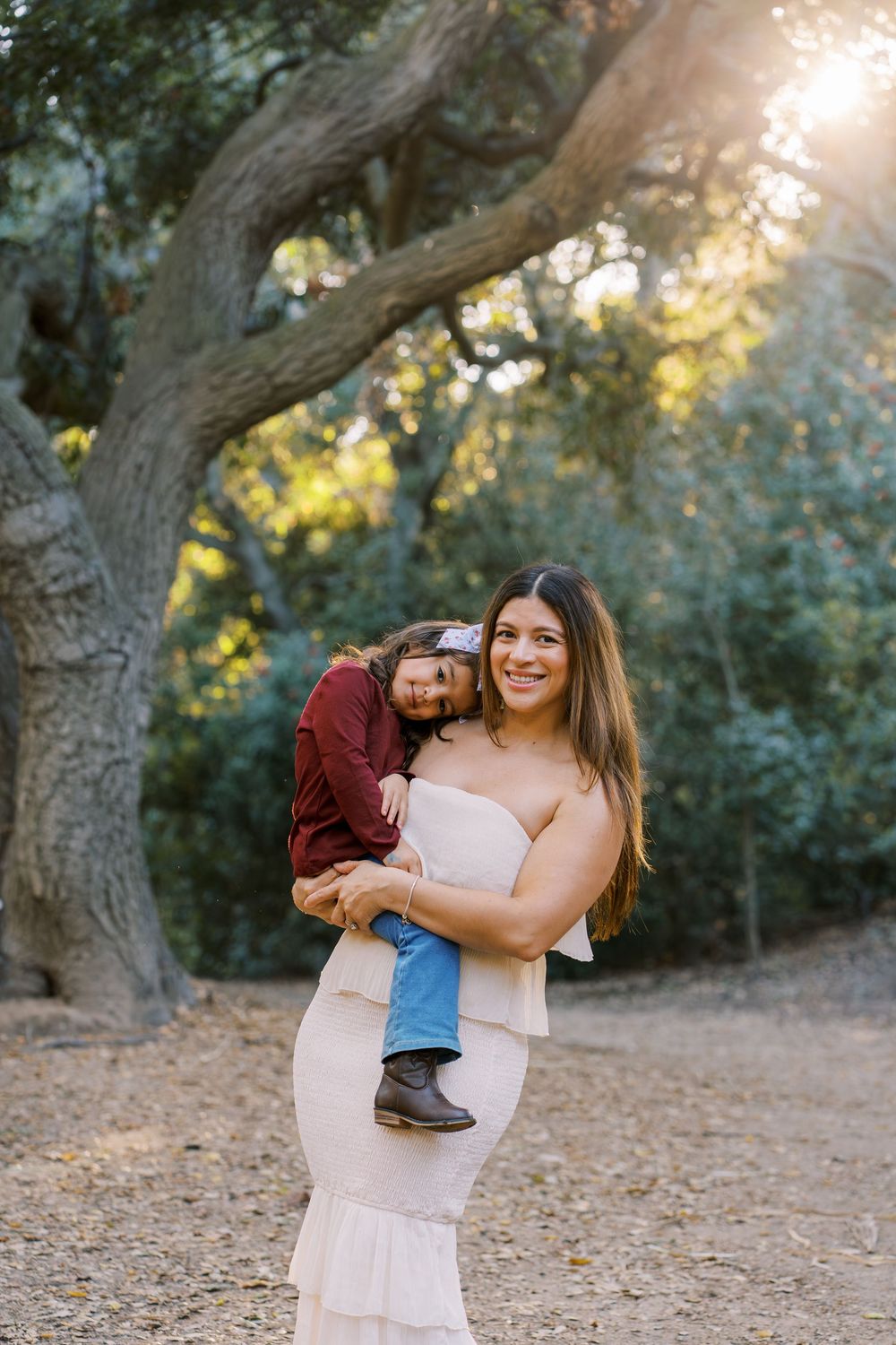 A parent and child share a joyful moment together under a large tree during golden hour in a natural outdoor setting.