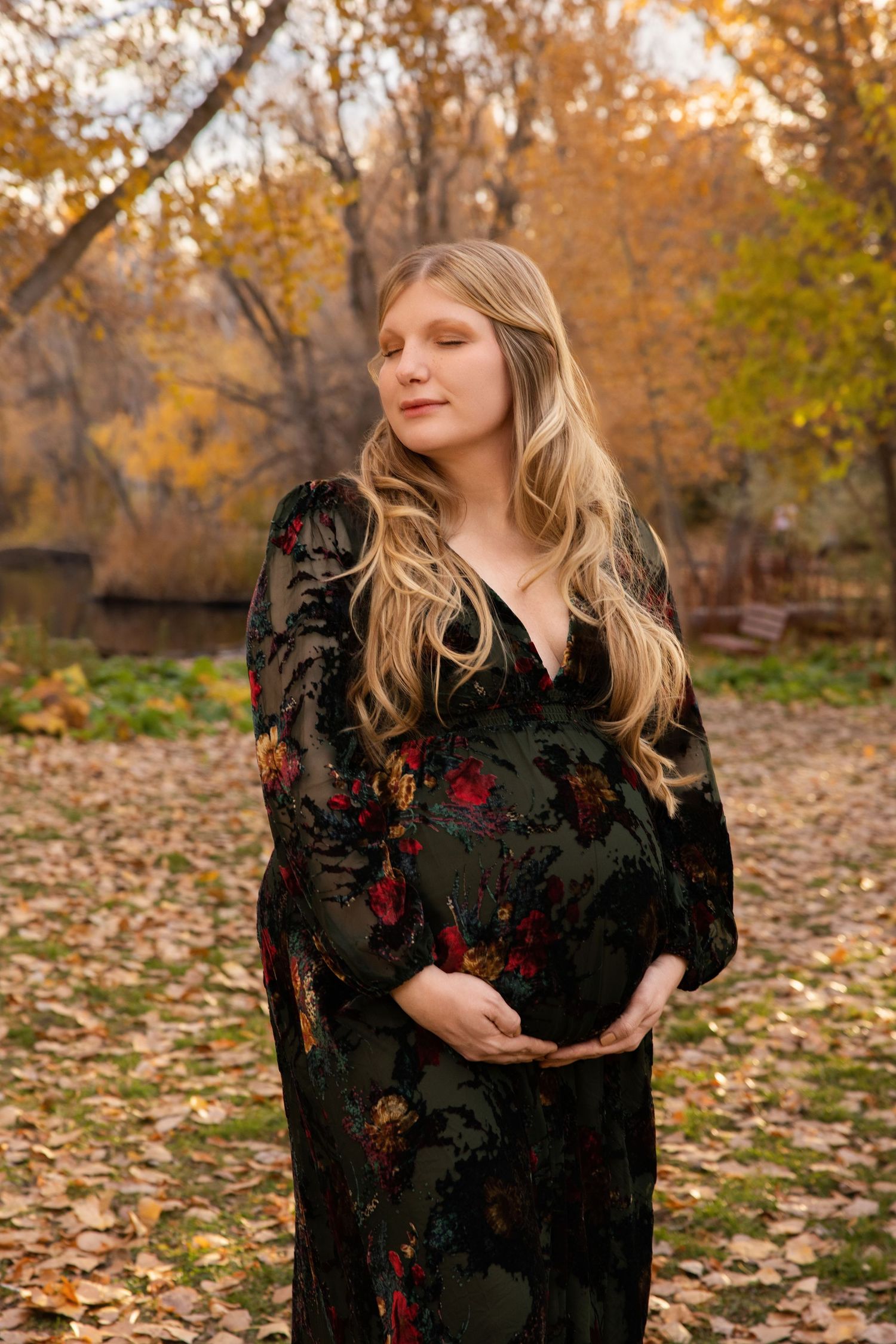 A person in a black floral dress poses gracefully among autumn leaves in a park during golden hour.