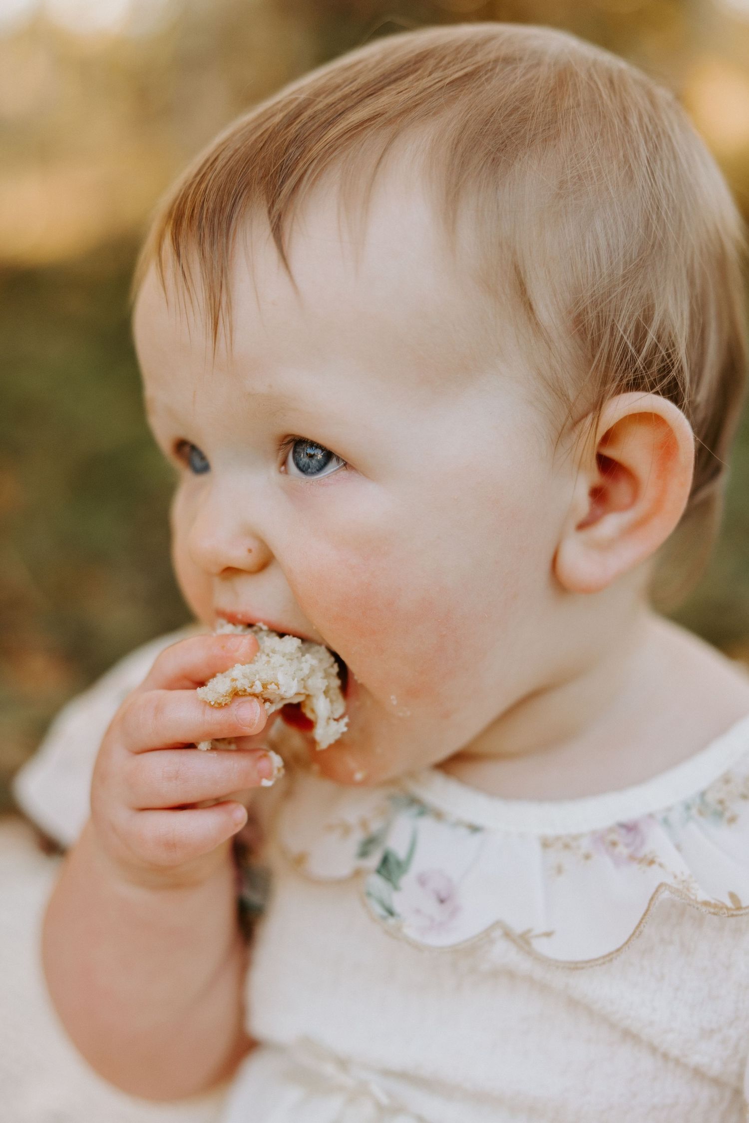 A small child with blue eyes wearing a floral dress takes a bite of food while sitting outdoors in warm lighting.