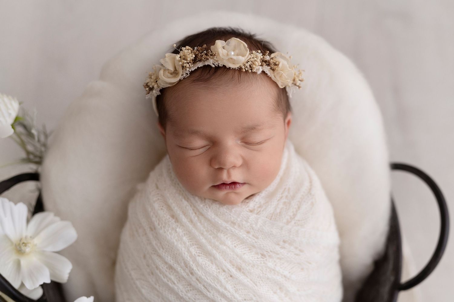 Sleeping newborn baby wrapped in soft white blankets with delicate white flowers and dark background.