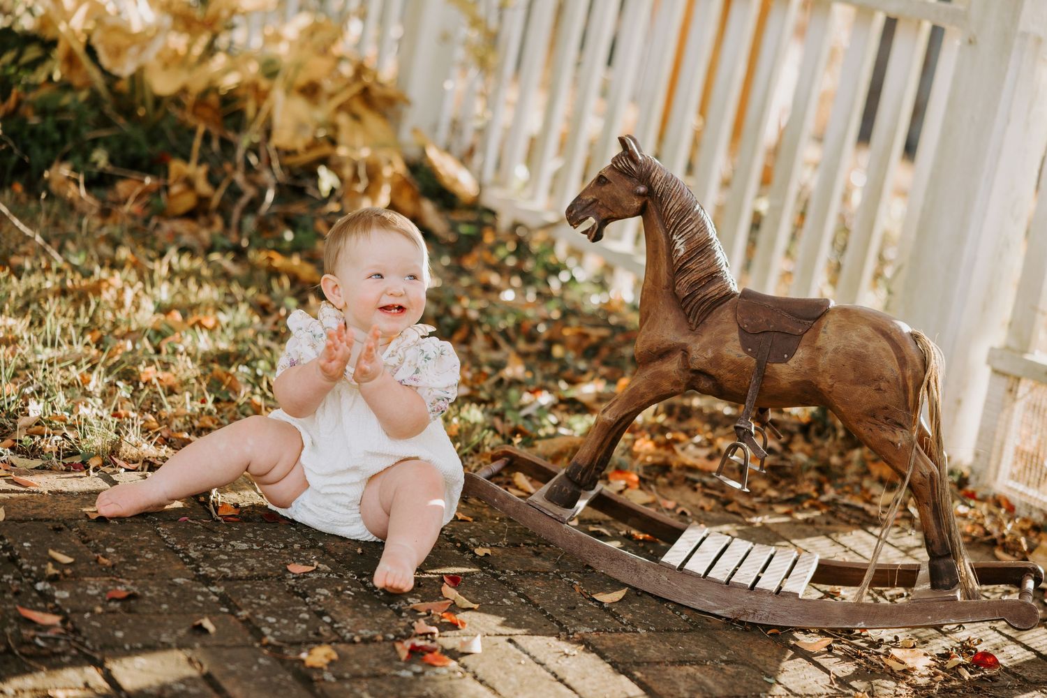 Baby in white dress explores and plays in garden setting with natural sunlight filtering through picket fence.