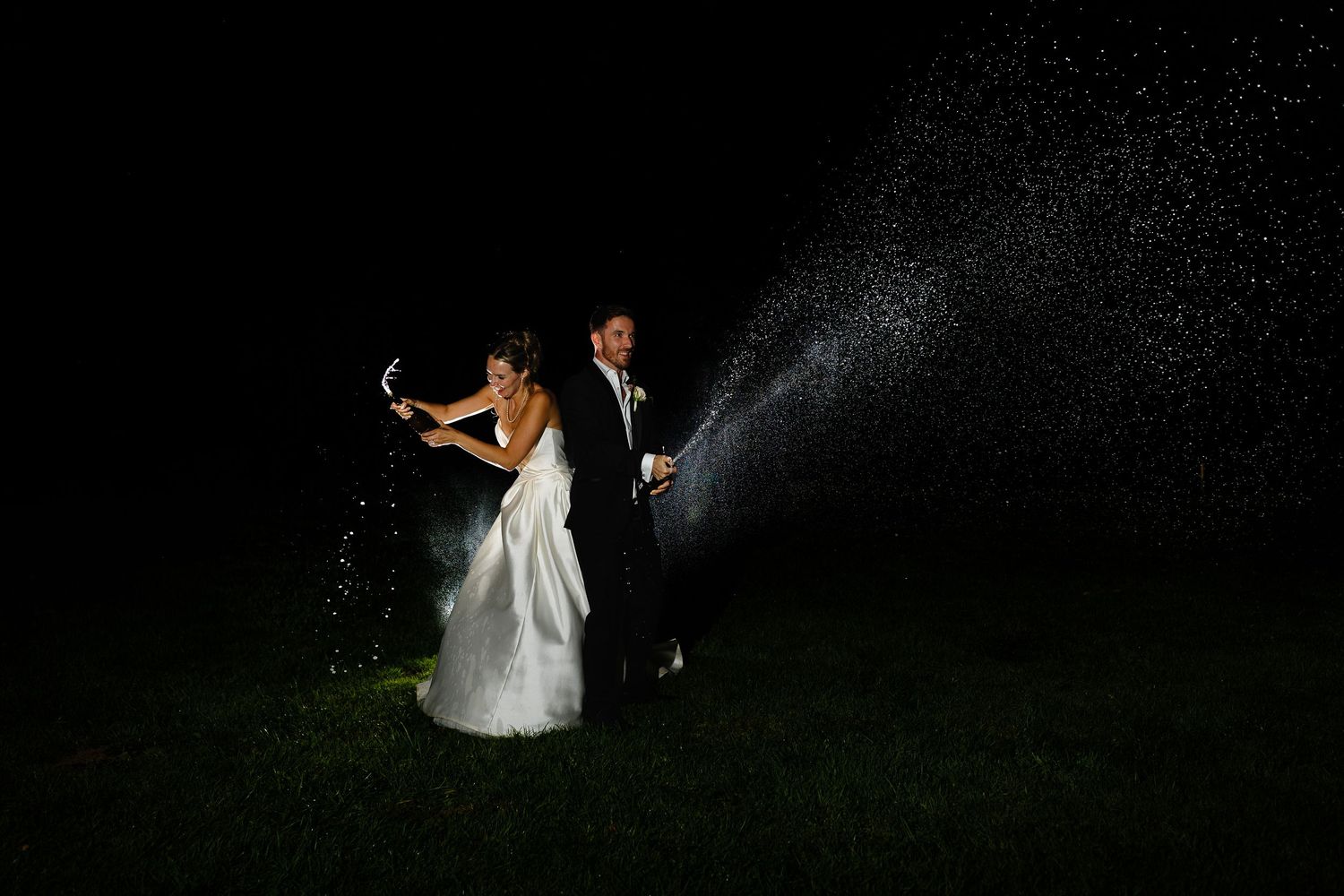 A couple in wedding attire dances on a dark lawn with a dramatic backlit spray of water creating a magical nighttime effect.