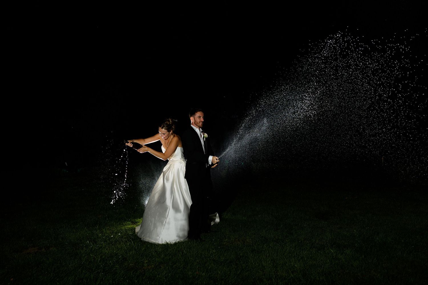 A wedding couple dances together under a spray of water in a dark outdoor setting creating a magical silhouette effect.