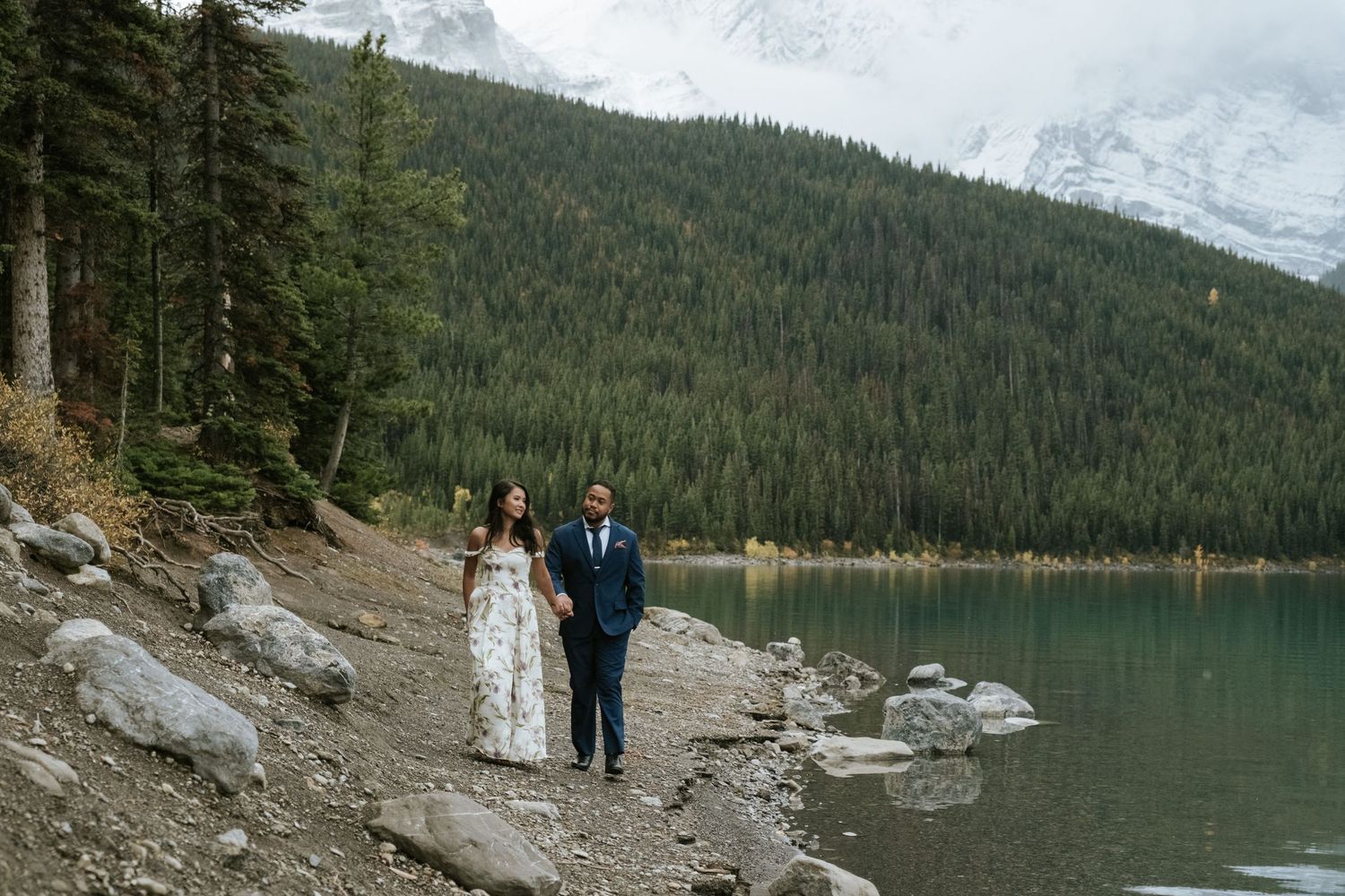 Two people in wedding attire walk along the rocky shore of a mountain lake surrounded by evergreen forest and peaks.