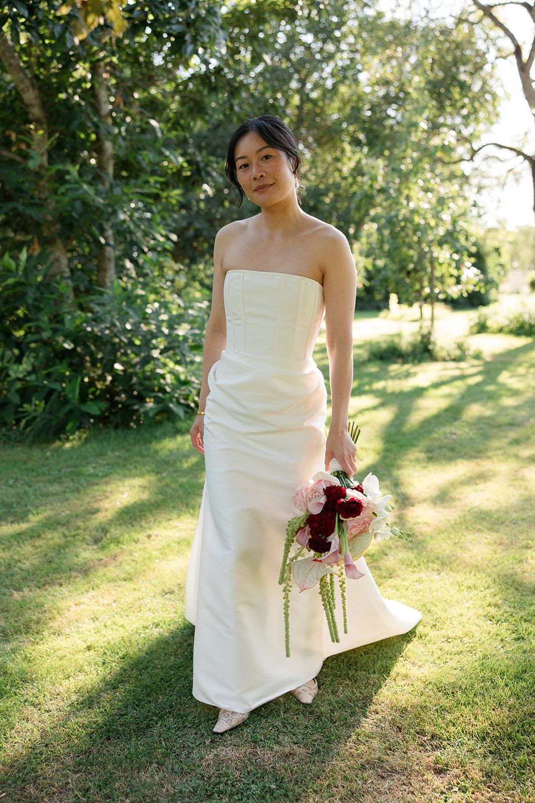 A bride in a white wedding dress poses gracefully in a sunlit garden setting holding a deep red bouquet.