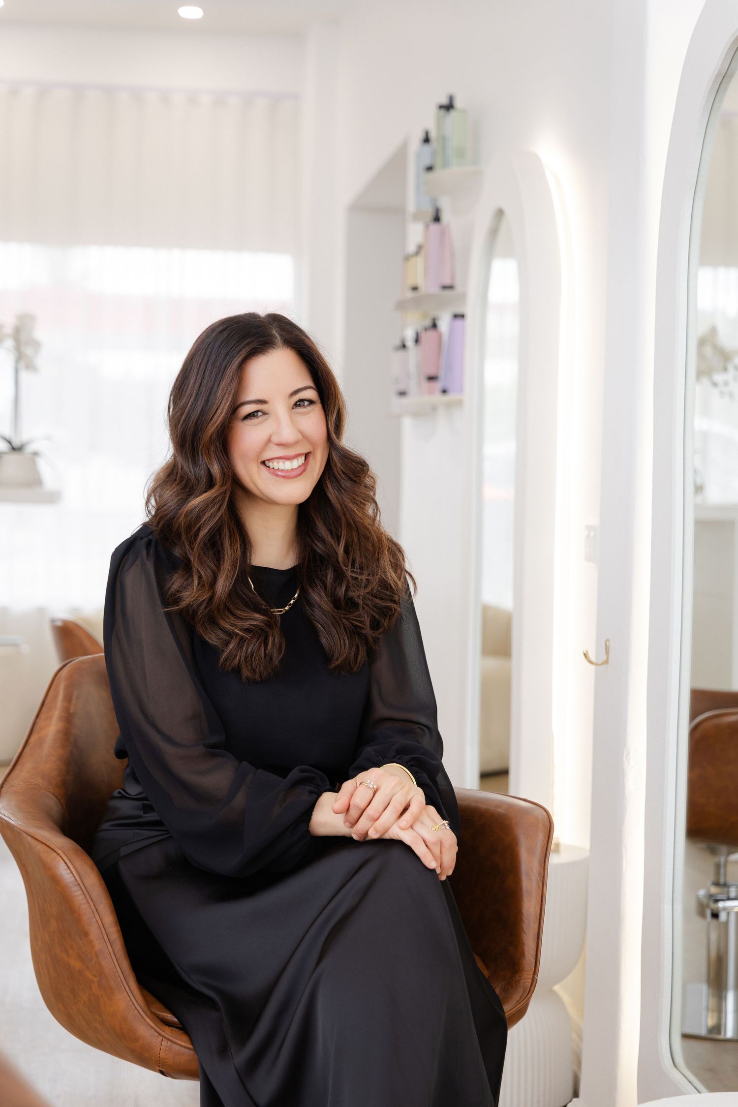 Professional lifestyle portrait of someone in a black turtleneck dress seated in a brown leather chair in a bright studio.