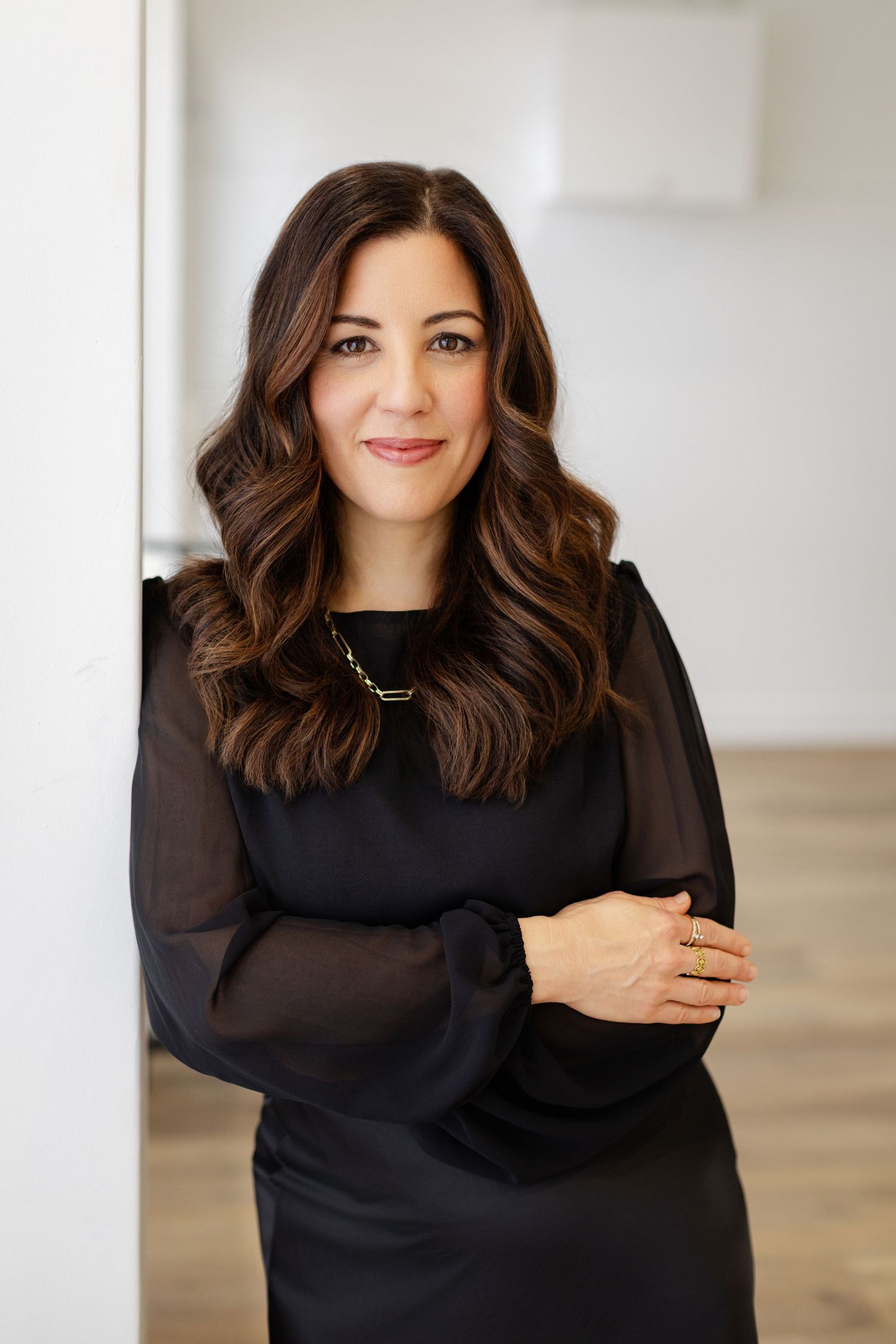 Professional portrait against white wall showing person in black long sleeve top with warm smile and wavy brown hair.