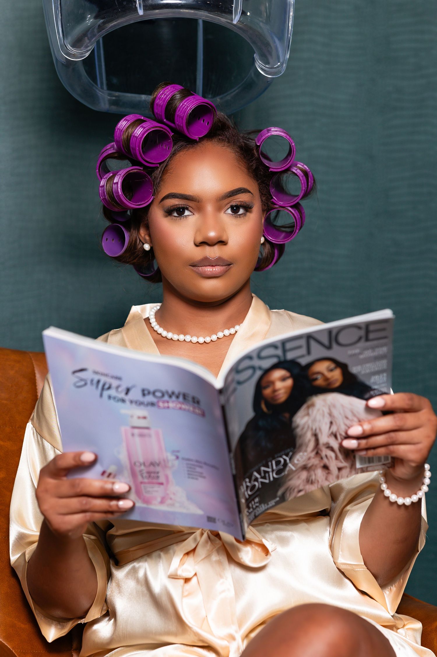 A person wearing hair rollers and a silk robe reads an Essence magazine while relaxing at home.