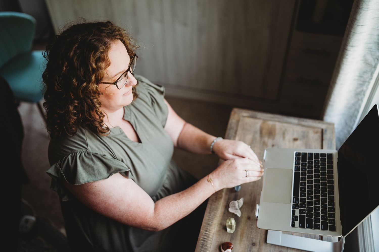 Person wearing glasses works on laptop computer while sitting at desk near window in natural light.