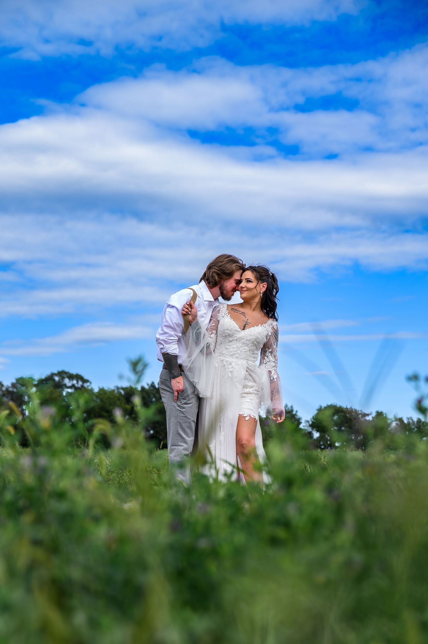 Romantic couple embracing in tall grass field under bright blue summer sky.