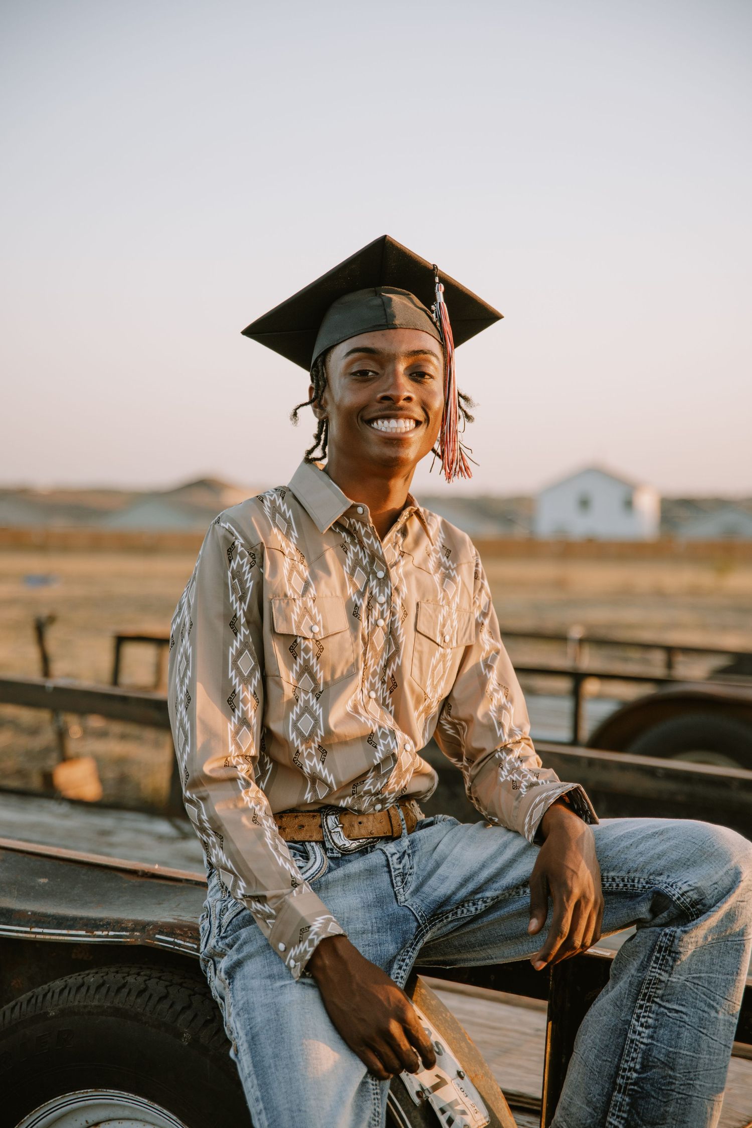 Graduate in a black cap wearing denim jeans and patterned shirt poses in desert landscape at sunset.