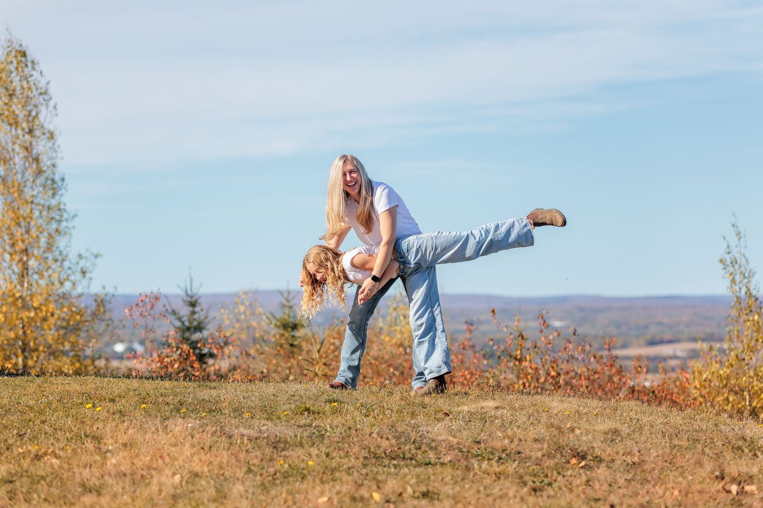 Two people enjoy a playful moment on a hilltop during autumn with mountains visible in the background.