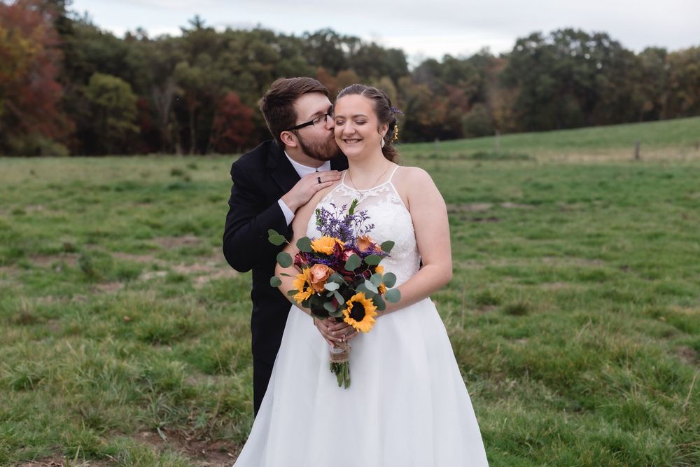A happy bride smiles as her husband kisses her on the cheek in a country field on their wedding day in Windham, New Hampshire.