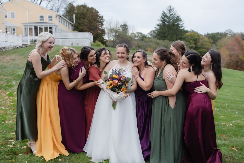 Bridesmaids hug a bride in a field outside historical Westbrook Inn in Derry, New Hampshire for bridal party photos. Concord wedding photos
