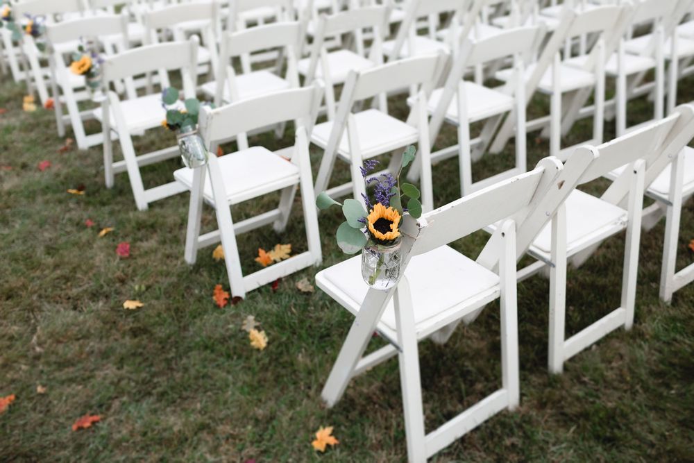 White folding chairs arranged on grass with scattered leaves creating a wedding ceremony aisle for a farm wedding in New Hampshire.