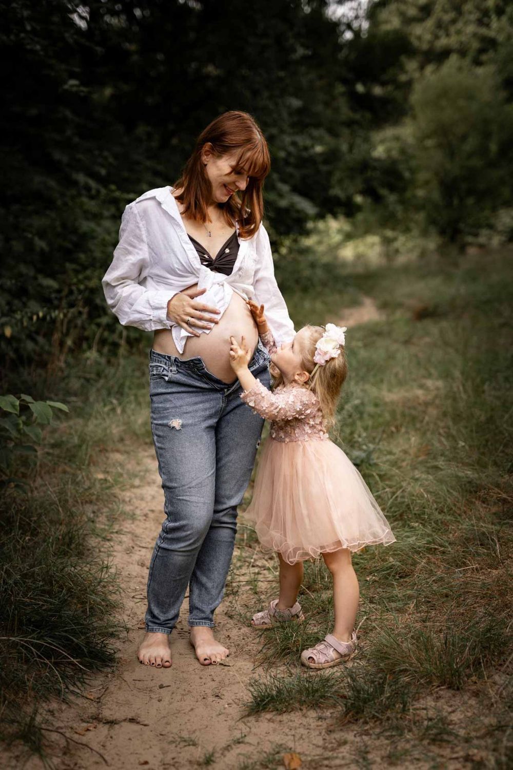 A pregnant woman in jeans and white shirt stands barefoot on a dirt path with a small child in a pink tulle dress.