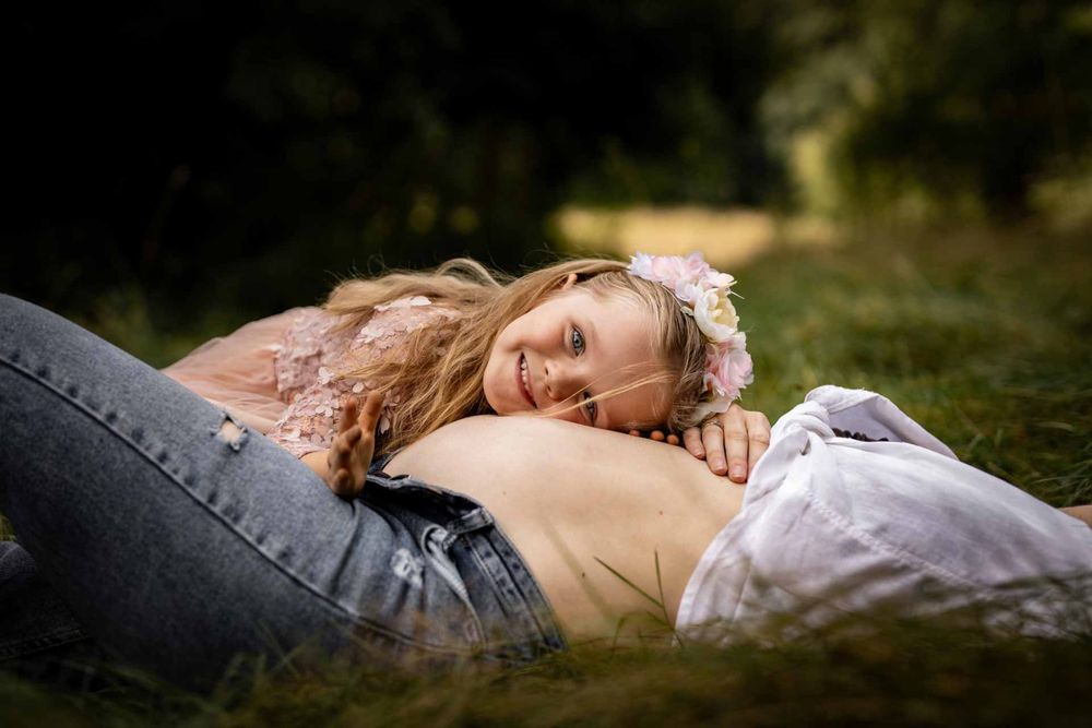 A person lies in grass wearing a white floral dress while surrounded by natural outdoor lighting and greenery.